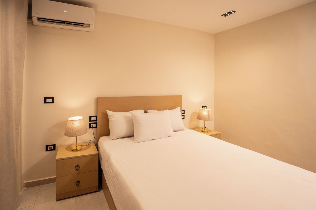 A neatly made bed with white linens, two bedside lamps, a wooden nightstand with drawers, and a wall-mounted air conditioner.
