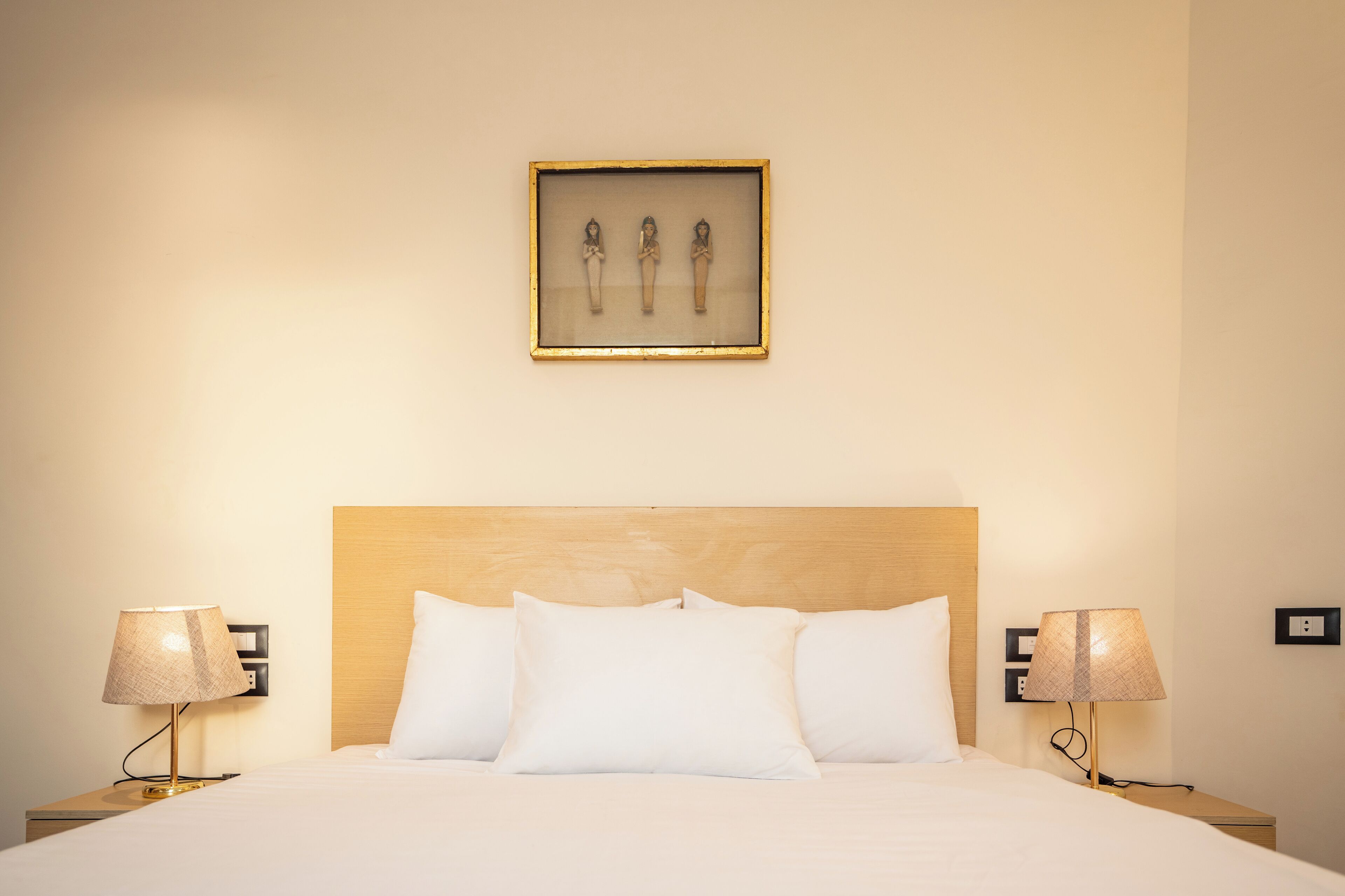 Quarto de hotel com cabeceira de madeira, roupa de cama branca, duas mesas de cabeceira com abajures e um quadro emoldurado sobre a cama.