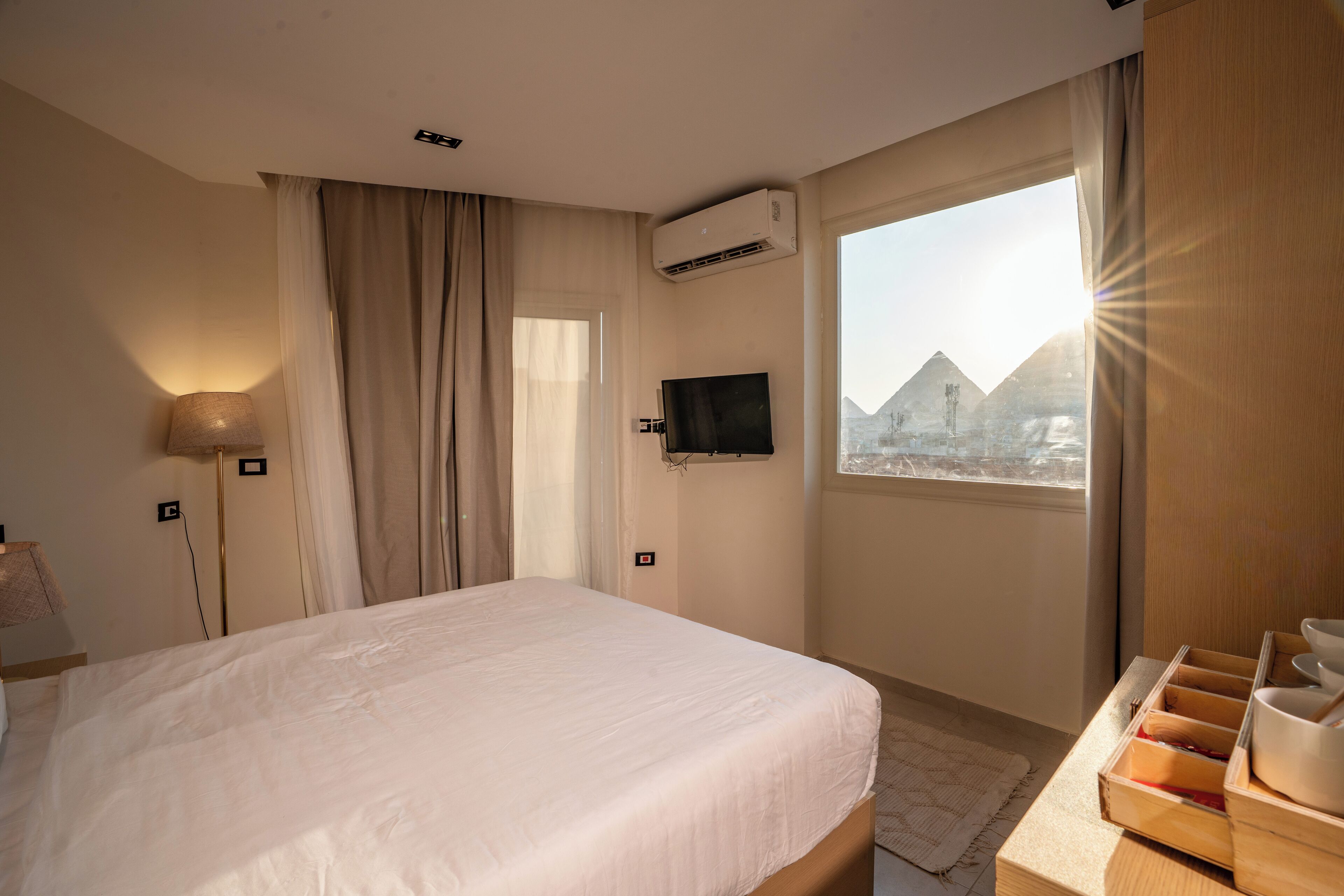 Une chambre d’hôtel comprenant un lit, un téléviseur fixé au mur, un appareil de climatisation, une fenêtre donnant sur les pyramides et une table d’appoint en bois avec des bols.