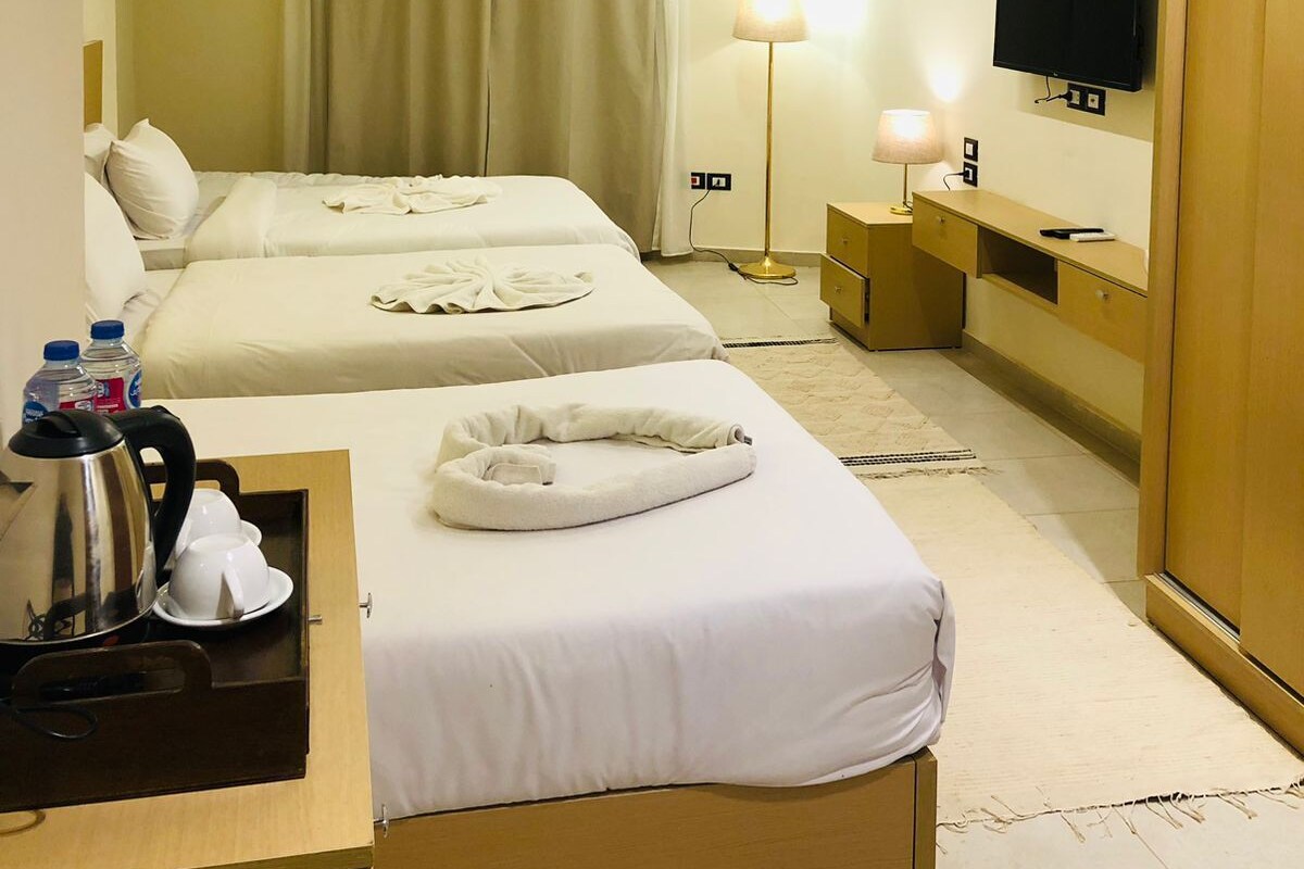 A hotel room with two beds, a desk with a kettle and cups, a bedside lamp, a TV, and a wooden wardrobe.