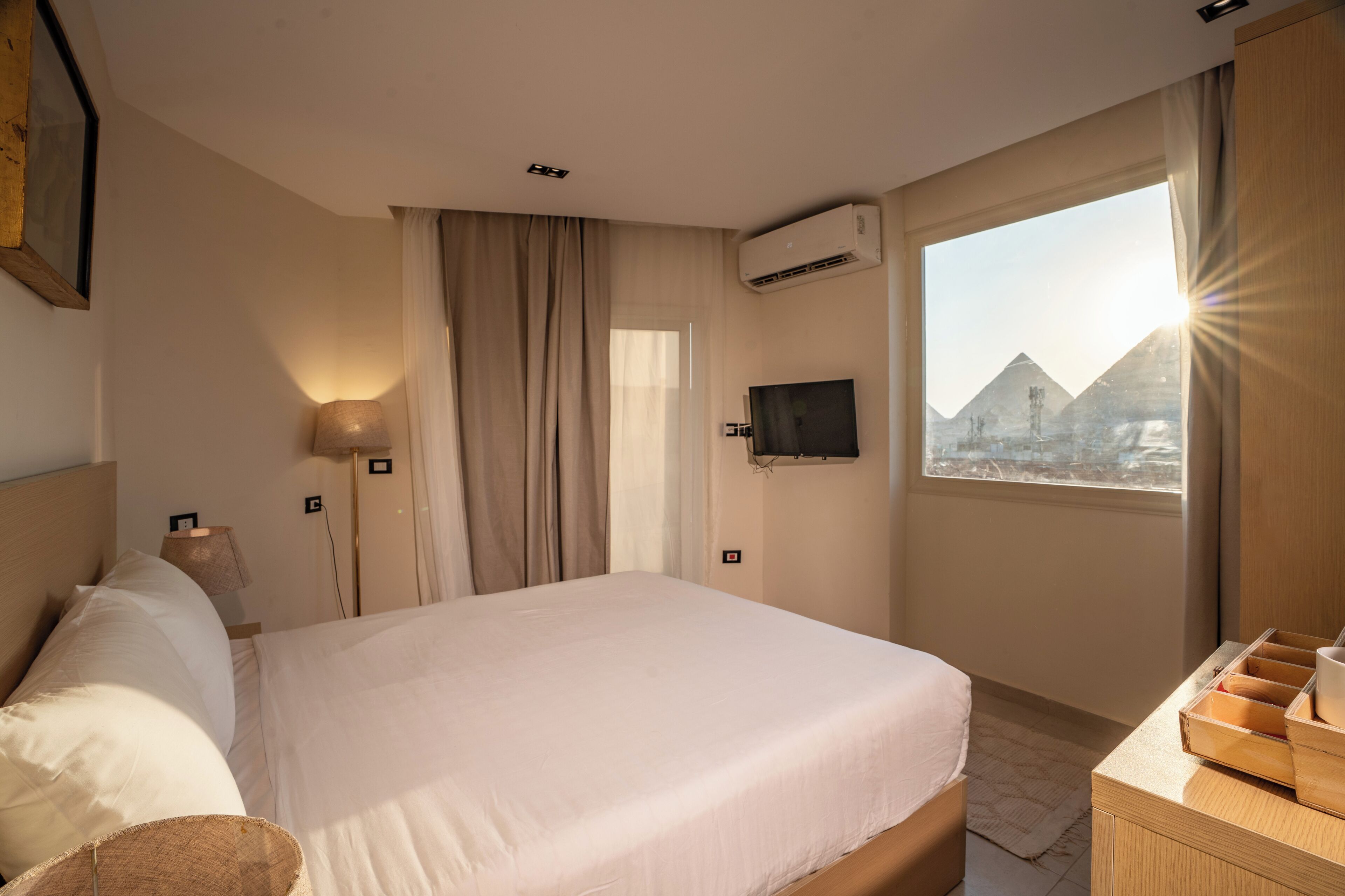 A hotel room with a large bed, a flat-screen TV mounted on the wall, a window with a view of pyramids, and a wooden dresser.
