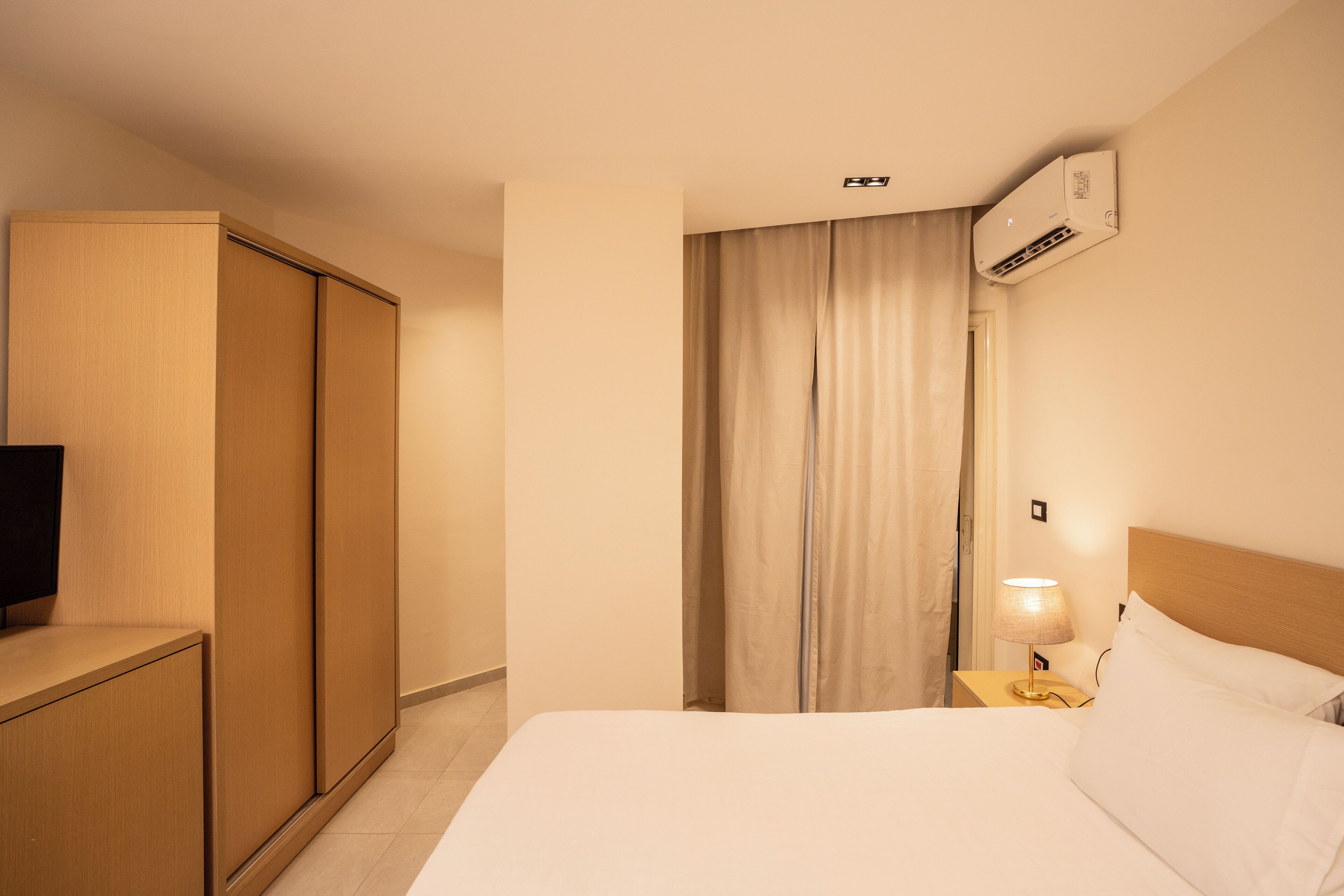 Habitación de hotel con cama, televisión, armario, aire acondicionado, mesita de noche con lámpara y cortina.