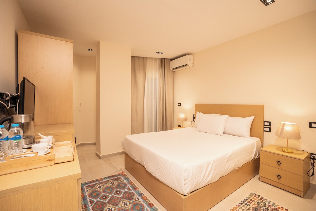 A hotel room with a bed, bedside table, lamp, and a desk with a coffee maker and cups.