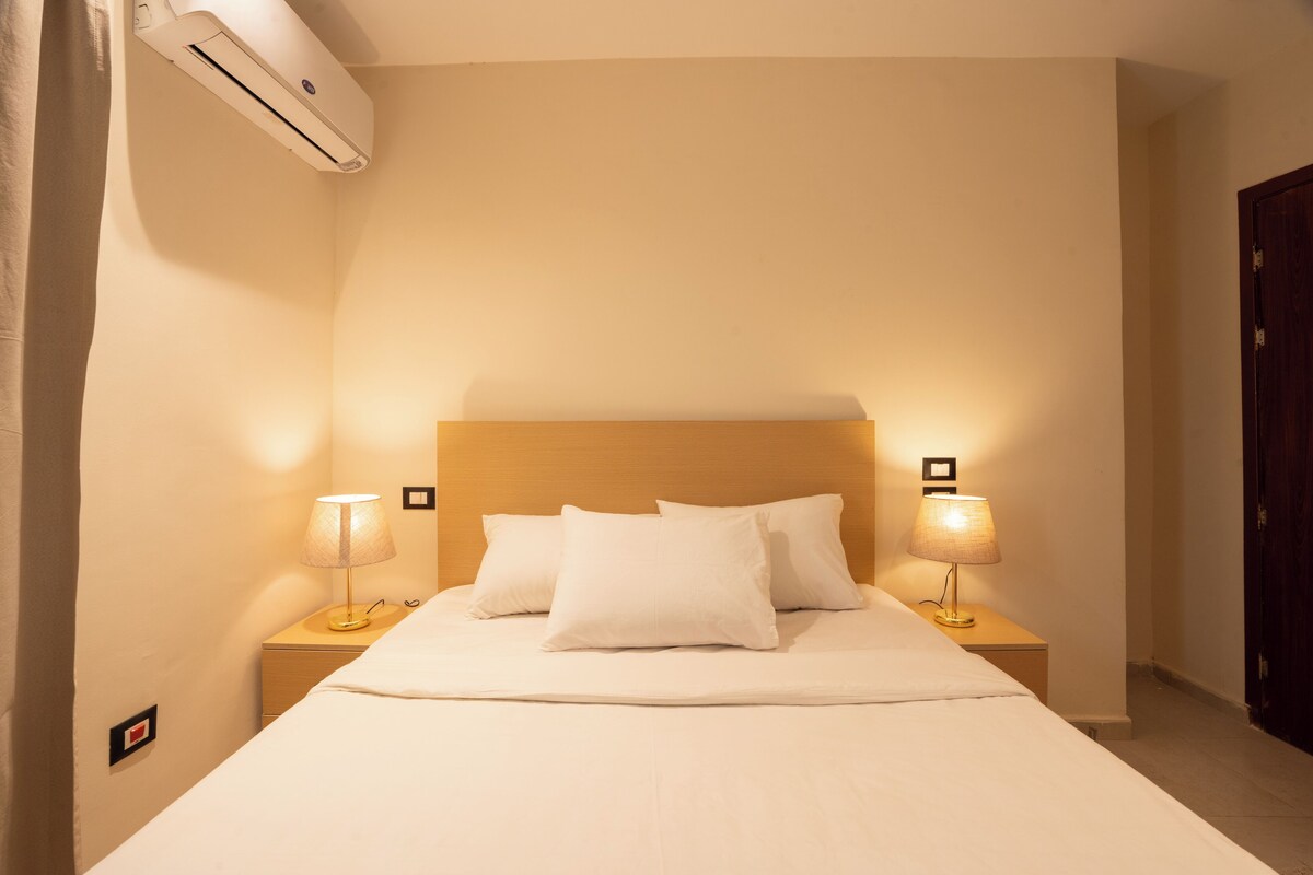 A neatly made bed with white linens, two bedside lamps, and an air conditioning unit on the wall.