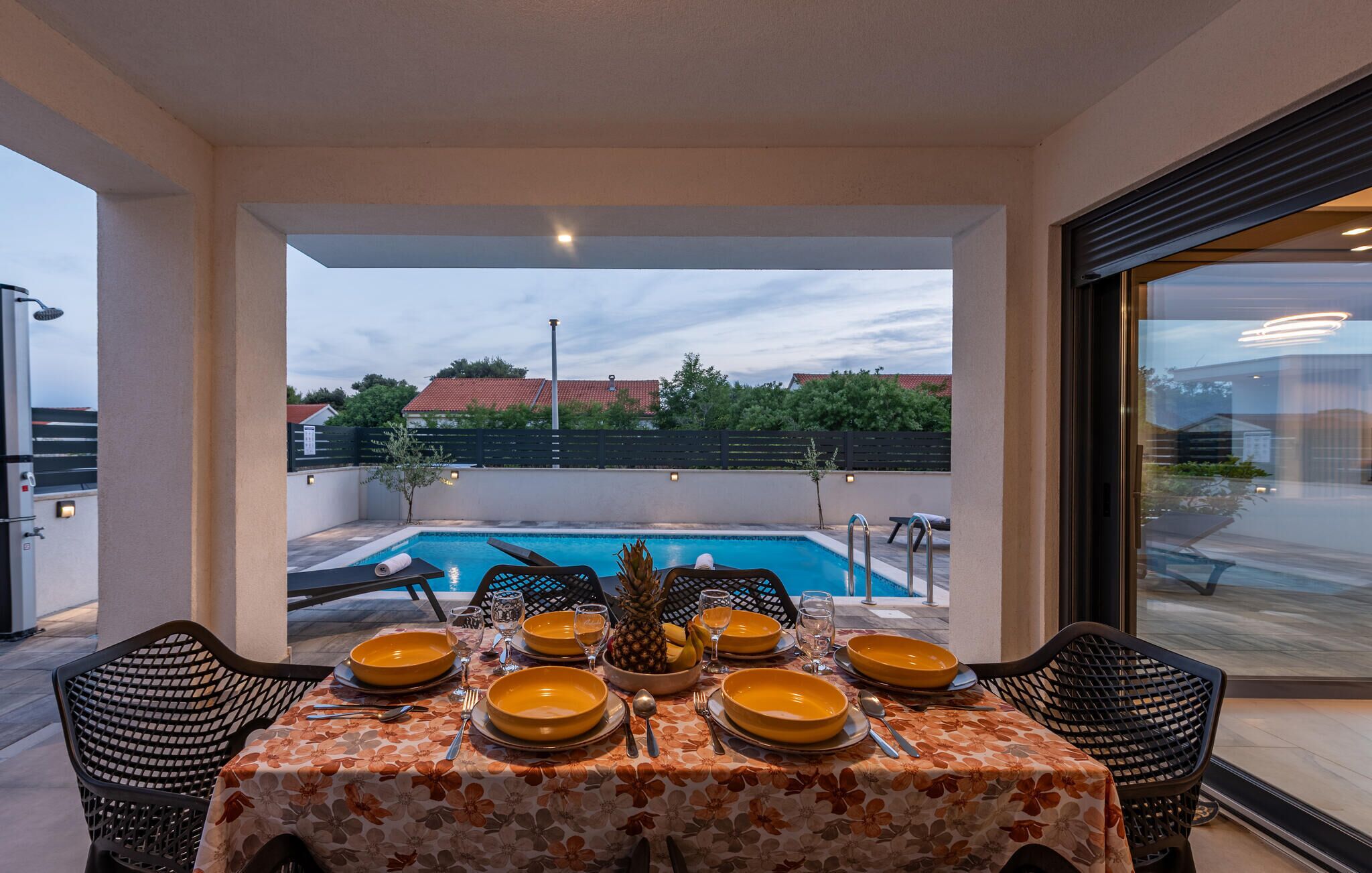 Comedor con una mesa dispuesta para cuatro, con vista a una piscina, y al fondo casas y árboles.
