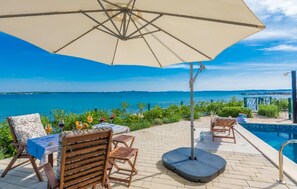A poolside area with a table, chairs, and an umbrella overlooking a body of water. - Cozy home in Privlaka (Privlaka)