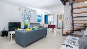 A modern living room with a grey sofa, a flat-screen TV, and a dining area with a red tablecloth. - Cozy home in Privlaka (Privlaka)