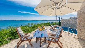 A terrace with a table, chairs, and a large umbrella overlooking a pool and the sea. - Cozy home in Privlaka (Privlaka)