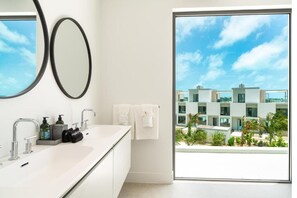 Une salle de bain moderne avec une grande fenêtre, un lavabo blanc, deux miroirs et une vue sur un quartier résidentiel.