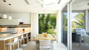 Eine moderne Küche mit einer zentralen Kochinsel, Barhocker und einem Esstisch mit Stühlen. Ein großes Schiebeglasfenster führt zu einer Terrasse mit einem Grill.