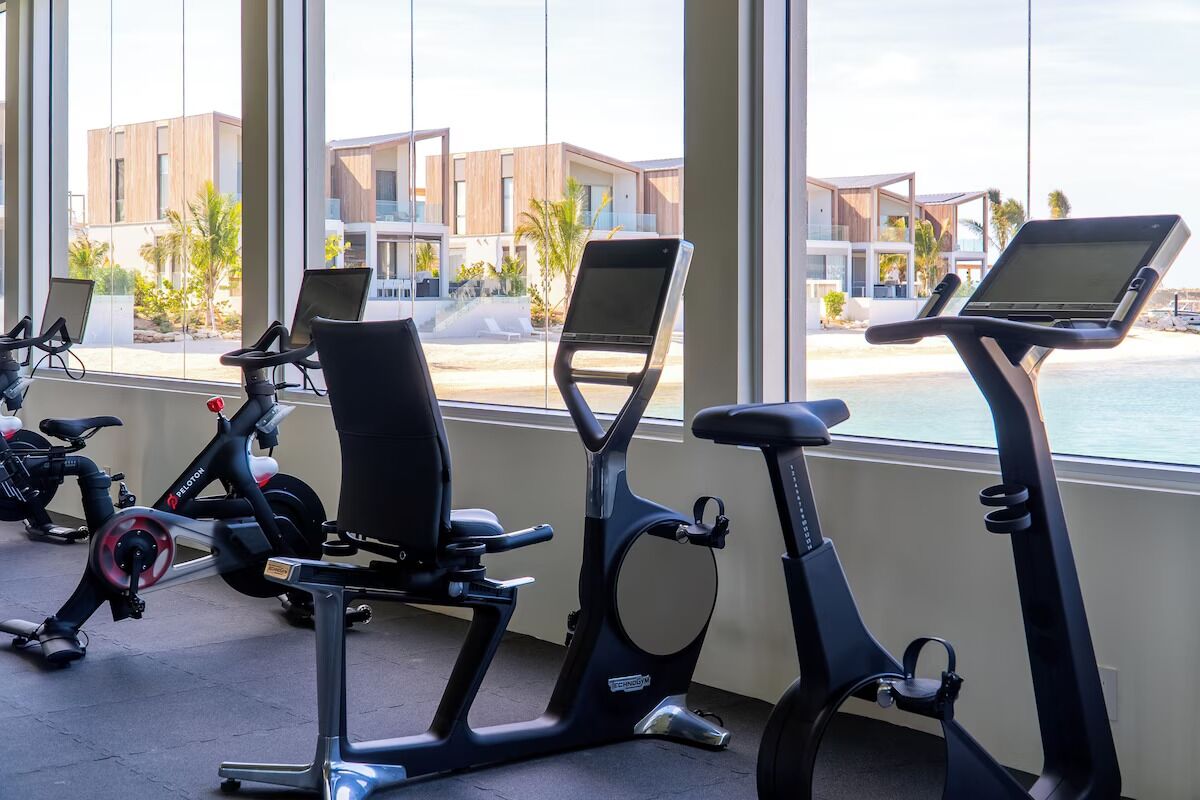 A row of stationary bikes with attached screens, positioned in a room with large windows overlooking a beach and modern buildings.