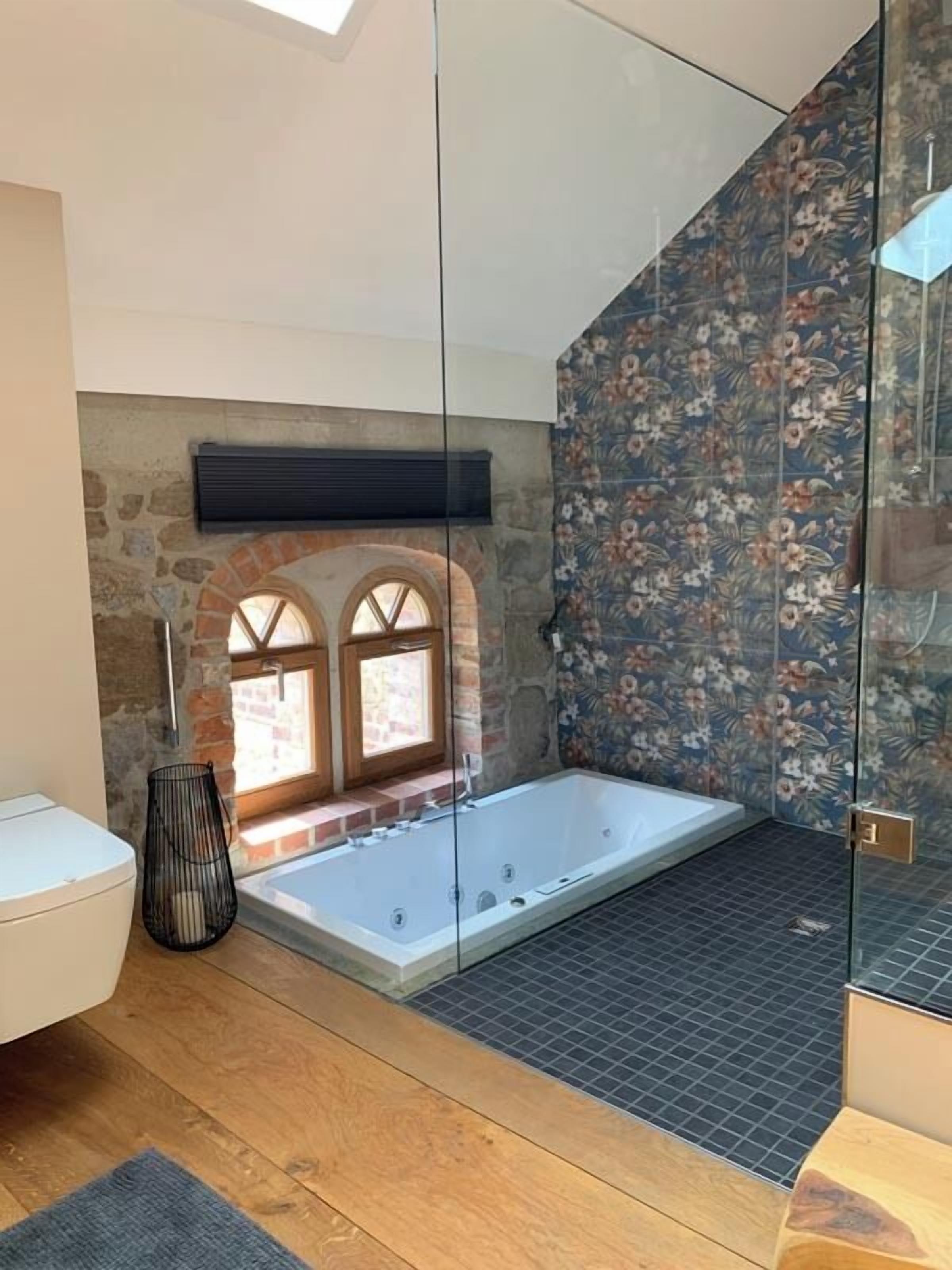 A modern bathroom with a freestanding bathtub, a glass shower enclosure, and a stone wall with arched windows.