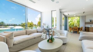 A modern living room with a large window overlooking a pool and ocean, a round coffee table with a plant, and a dining area with wooden furniture. - Turks Adventure | Kayak, Snorkel & Explore Turks’ Ocean Lagoon | Private Pool (Long Bay Hills)