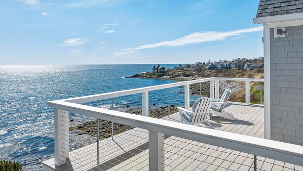 A coastal balcony with a view of the sea, a white railing, and two Adirondack chairs.
