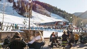 An outdoor seating area with chairs and tables, a ski lift in the background, and a building with signage. - Ski Chalet Beaver Creek - Ski In/Ski Out, Hot Tub (Beaver Creek)