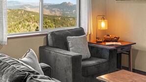 Una habitación acogedora con un sofá gris, una mesita con una lámpara y vistas a un paisaje montañoso a través de un ventanal grande.