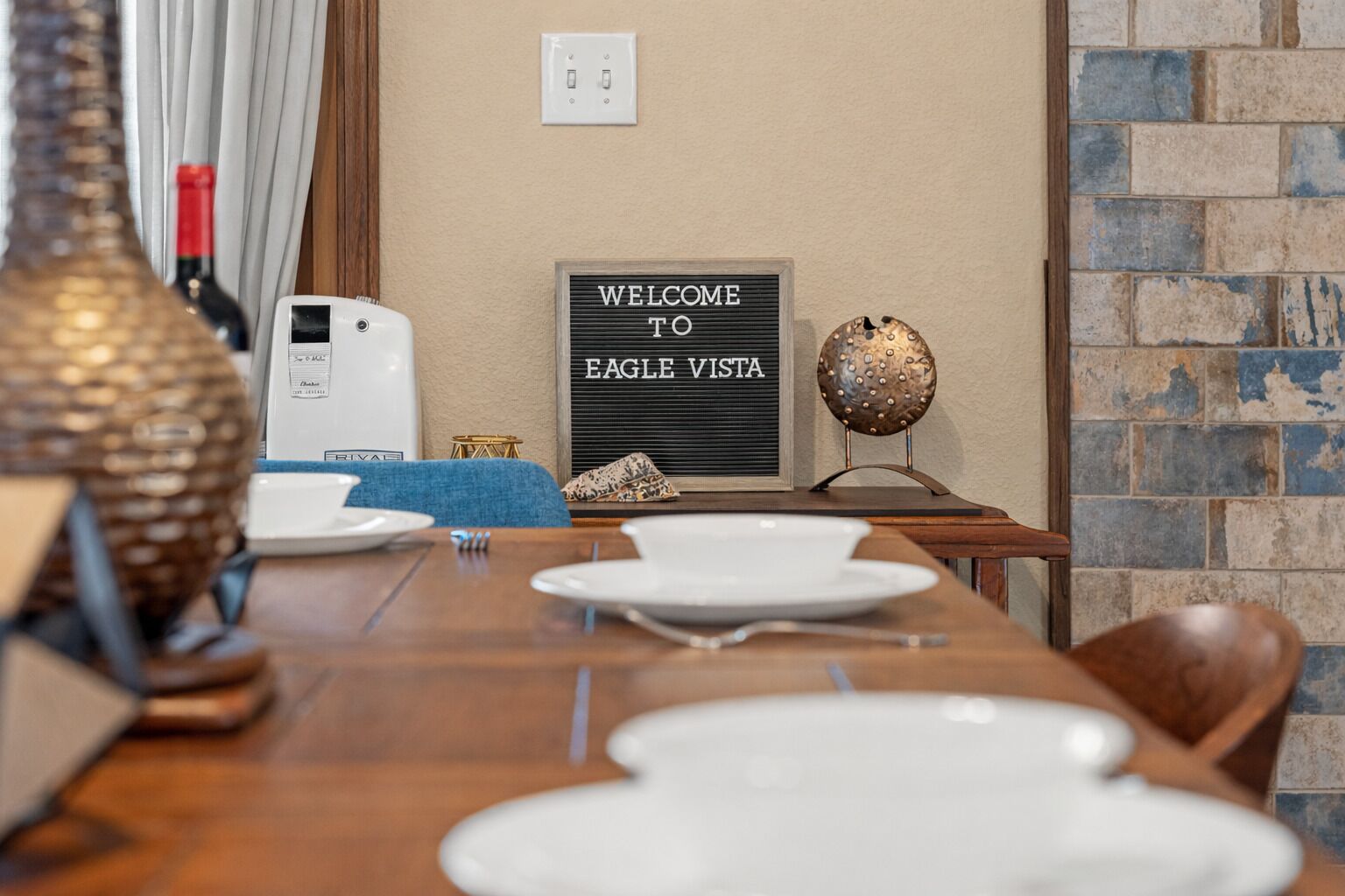 Zona pranzo con tavolo di legno apparecchiato per quattro persone, un cartello con scritto 'Welcome to Eagle Vista', una sfera metallica decorativa e un elettrodomestico bianco.