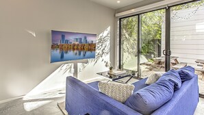 A modern living room with a large window, a blue sofa, and a cityscape painting on the wall. - New 2 BDRM Home I Mins to DT Walk to Shops & More (Austin)