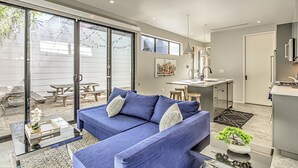 A modern living room with a blue sofa, a glass coffee table, and a kitchen area in the background. - New 2 BDRM Home I Mins to DT Walk to Shops & More (Austin)