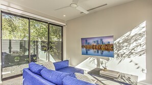 A modern living room with a large window, a blue sofa, a flat-screen TV, and a cityscape painting. - New 2 BDRM Home I Mins to DT Walk to Shops & More (Austin)