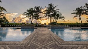 Uma área de piscina com um caminho de azulejos, espreguiçadeiras, palmeiras e vista para o pôr do sol.