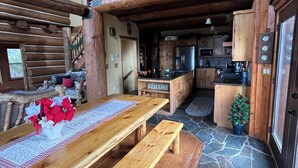 Interior rústico de una cabaña con una mesa de comedor de madera, bancos, una zona de cocina y un balcón con barandilla.