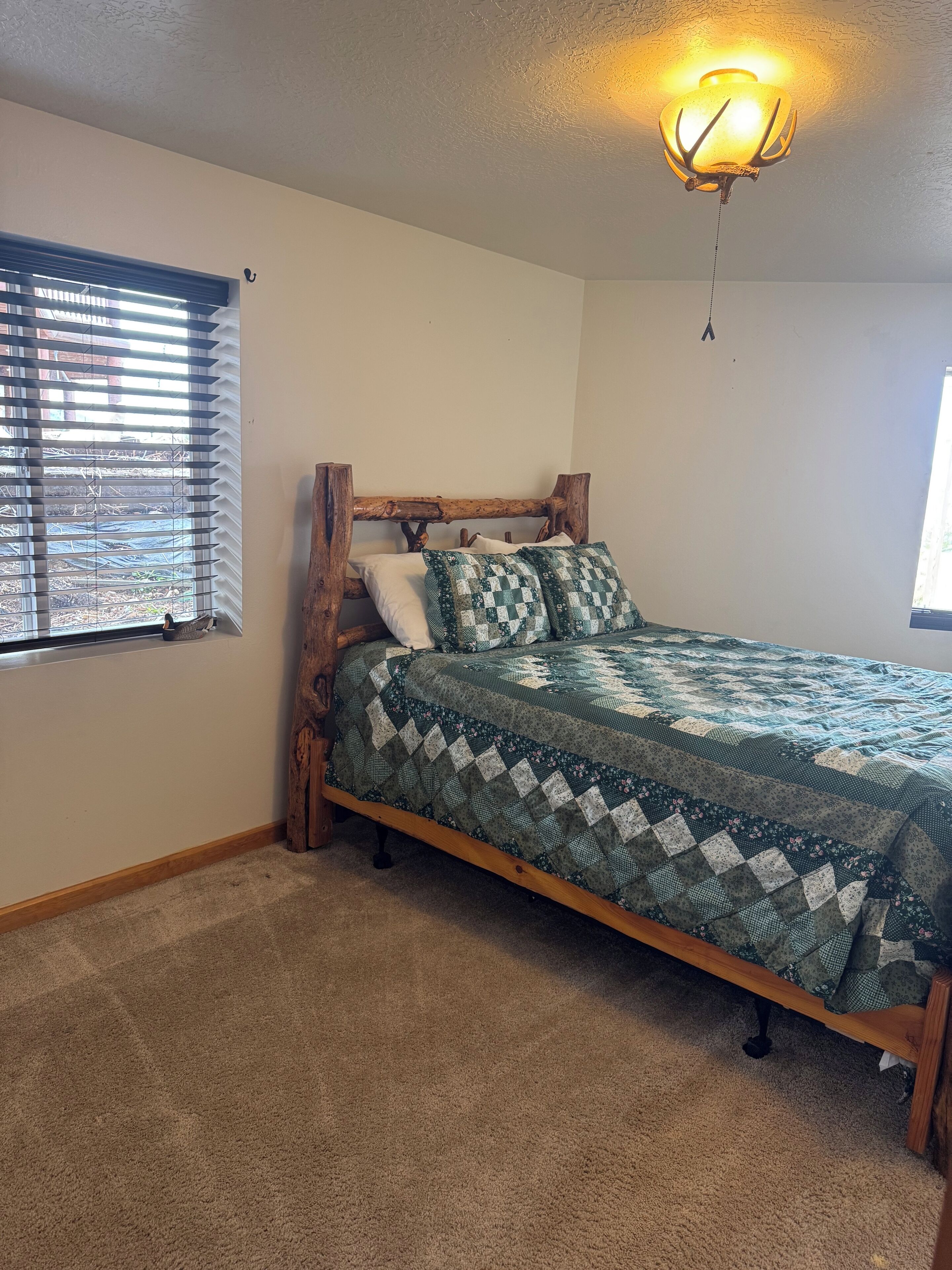 A bedroom with a wooden bed frame, a quilted bedspread, a ceiling light fixture, and a window with blinds.