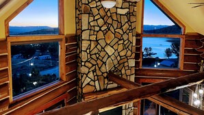 Una habitación con pared de piedra, vigas de madera y grandes ventanales que ofrecen vistas al paisaje.
