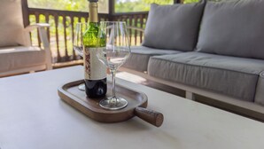 Una botella de vino y dos copas sobre una bandeja de madera en una mesa, con un sofá y una vista de árboles al fondo.