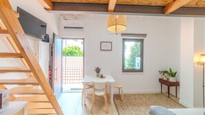 Een moderne, goed verlichte kamer met een houten trap, een eettafel met stoelen, een klein bijzettafeltje met een plant en een groot raam dat natuurlijk licht binnenlaat.