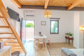 Une pièce moderne et bien éclairée, avec un escalier en bois, une table à manger entourée de chaises, une petite table d’appoint agrémentée d’une plante et une grande fenêtre laissant entrer la lumière naturelle.