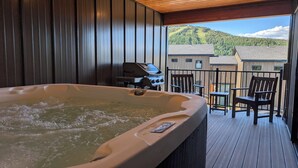 Un bain à remous sur une terrasse en bois, avec vue sur une montagne et des maisons.