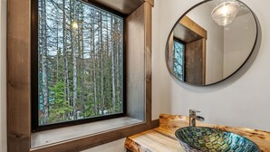 Une salle de bain avec un meuble-lavabo en bois, un miroir rond et une fenêtre donnant sur une forêt.