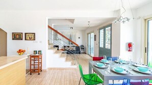 A modern kitchen with a wooden counter, a fruit bowl, and a cart. There is a dining area with a table set for four, chairs, and a staircase leading to another room.