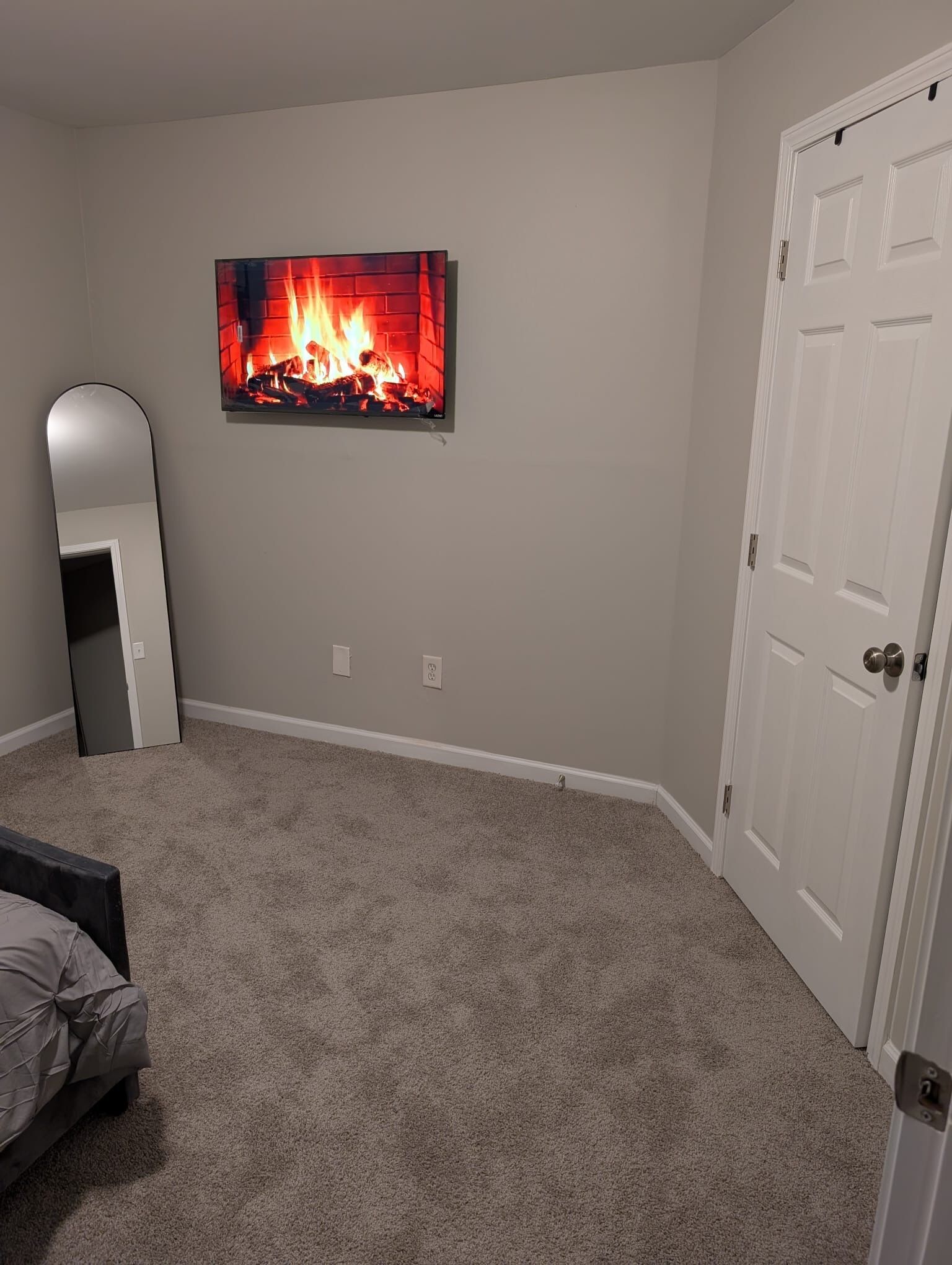 A bedroom with a carpeted floor, a bed, a wall-mounted TV displaying a fireplace, and a white door.
