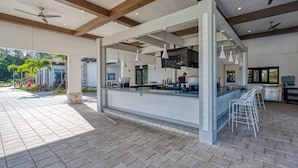 Uma área de bar externa com balcão, banquetas, ventiladores de teto, luminárias pendentes e vista do jardim ao redor.