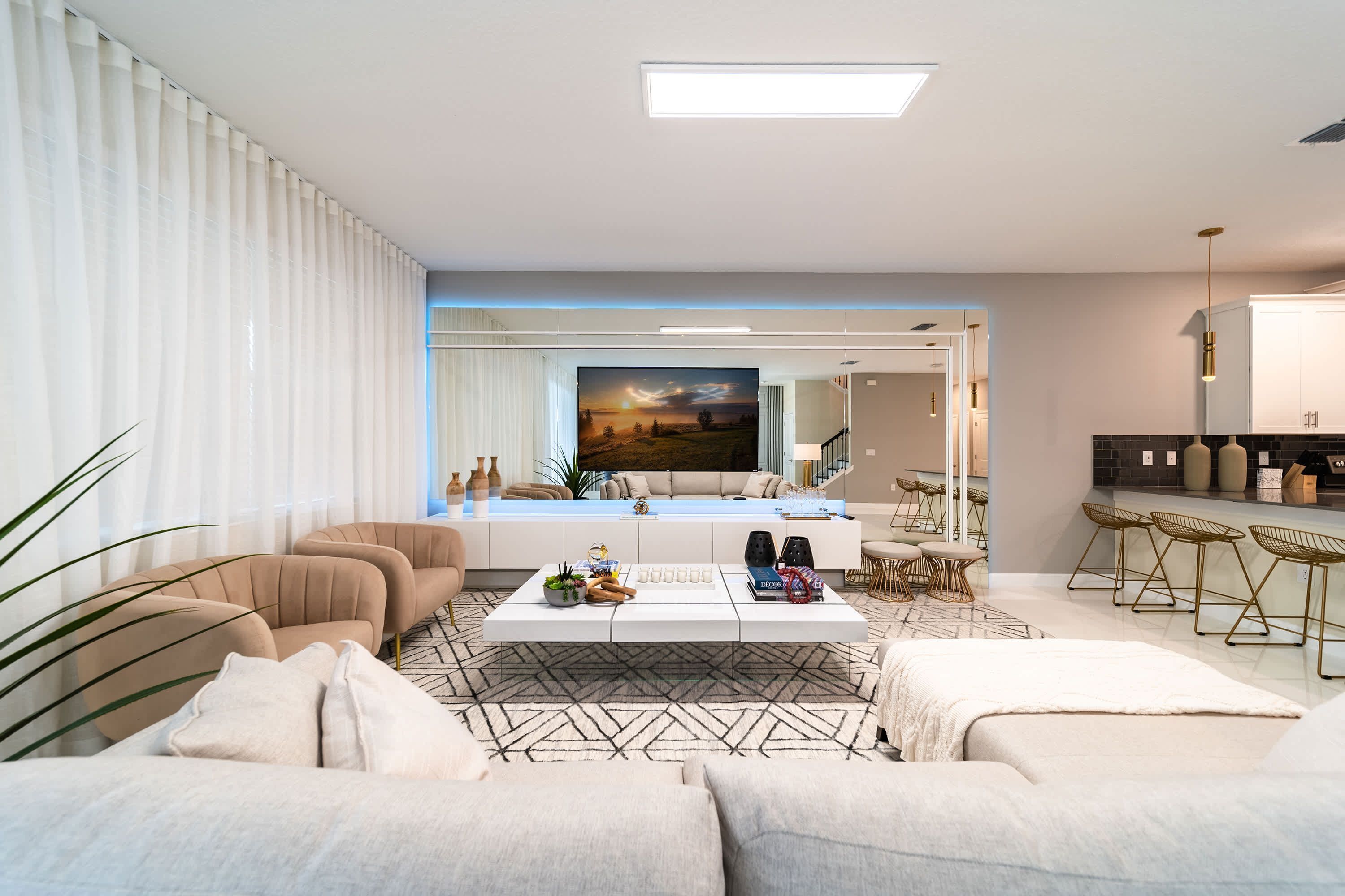 Een moderne woonkamer met een bank, salontafel en stoelen. Er is een groot televisiescherm zichtbaar en een keuken in de achtergrond.