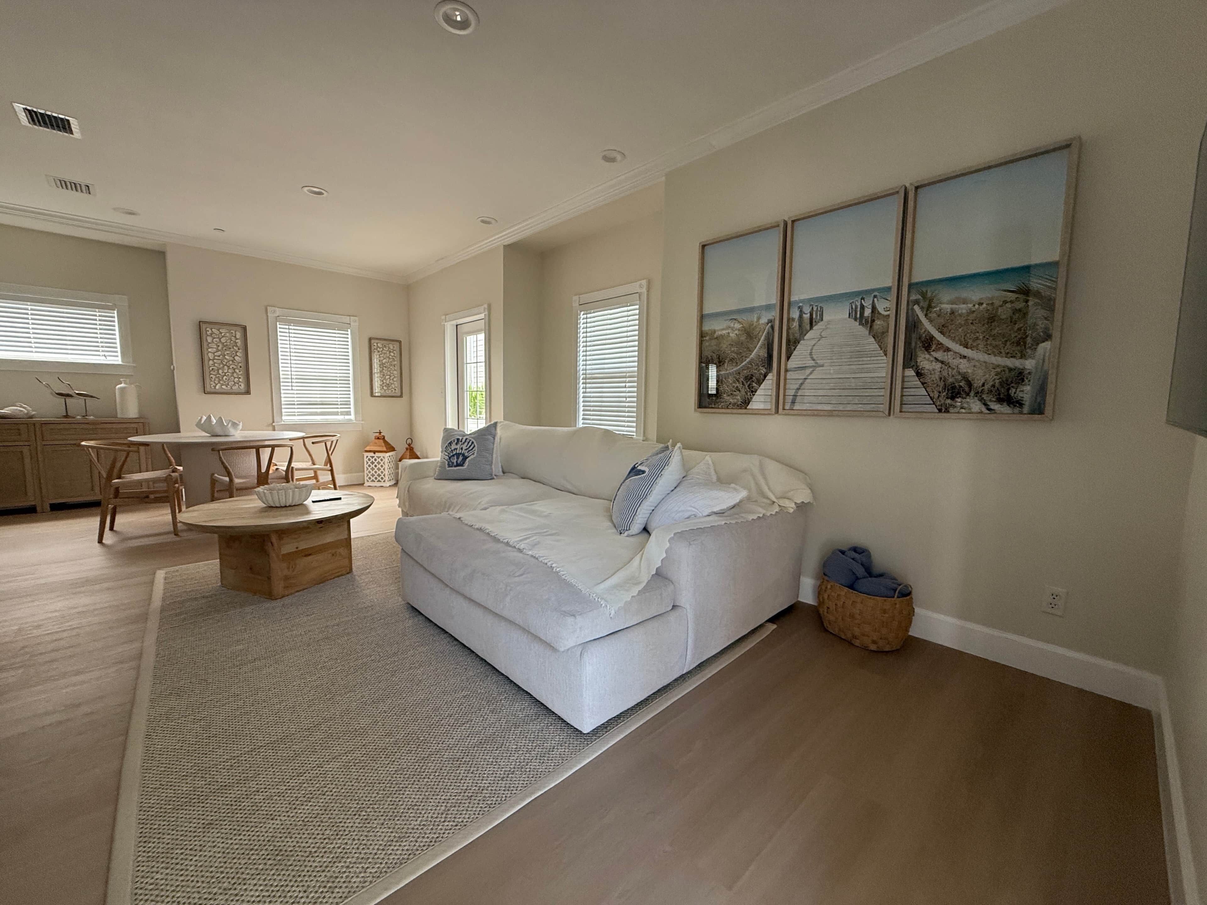 A spacious living room with a white sofa, a wooden coffee table, and a dining area with a white table and chairs. There are large windows with blinds, a kitchen in the background, and a beach-themed artwork on the wall.