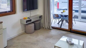 Ein modernes Zimmer mit Flachbildfernseher, einem kleinen Tisch mit einer Schachtel, einem Fenster mit Jalousien und Blick ins Freie.