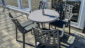 Ein runder Gartentisch mit vier Stühlen, jeweils mit Rücklehnen im Gittermuster. Auf dem Tisch steht ein kleiner Blumenstrauß mit violetten Blüten.
