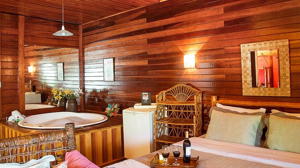 A wooden cabin-style room with a large bed, a wooden chair, a small table with a bottle and glasses, and a white refrigerator.