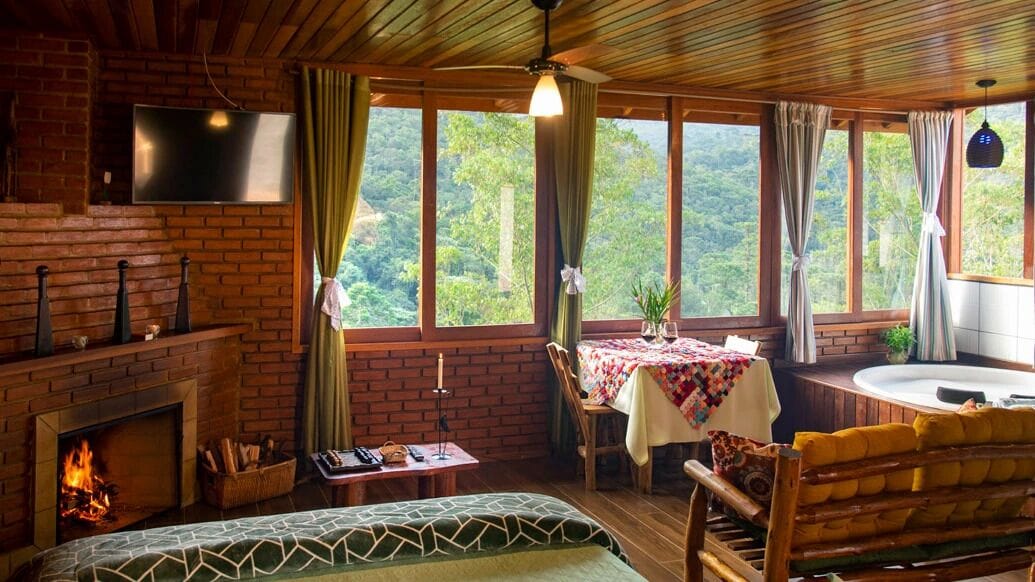 A cozy room with a fireplace, a dining table with a tablecloth, a sofa, and a view of the forest.