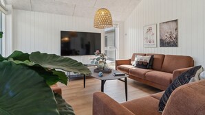 Uma sala de estar com um sofá marrom, uma mesa de centro e uma televisão. Há uma planta grande em primeiro plano e um lustre pendurado sobre o sofá.