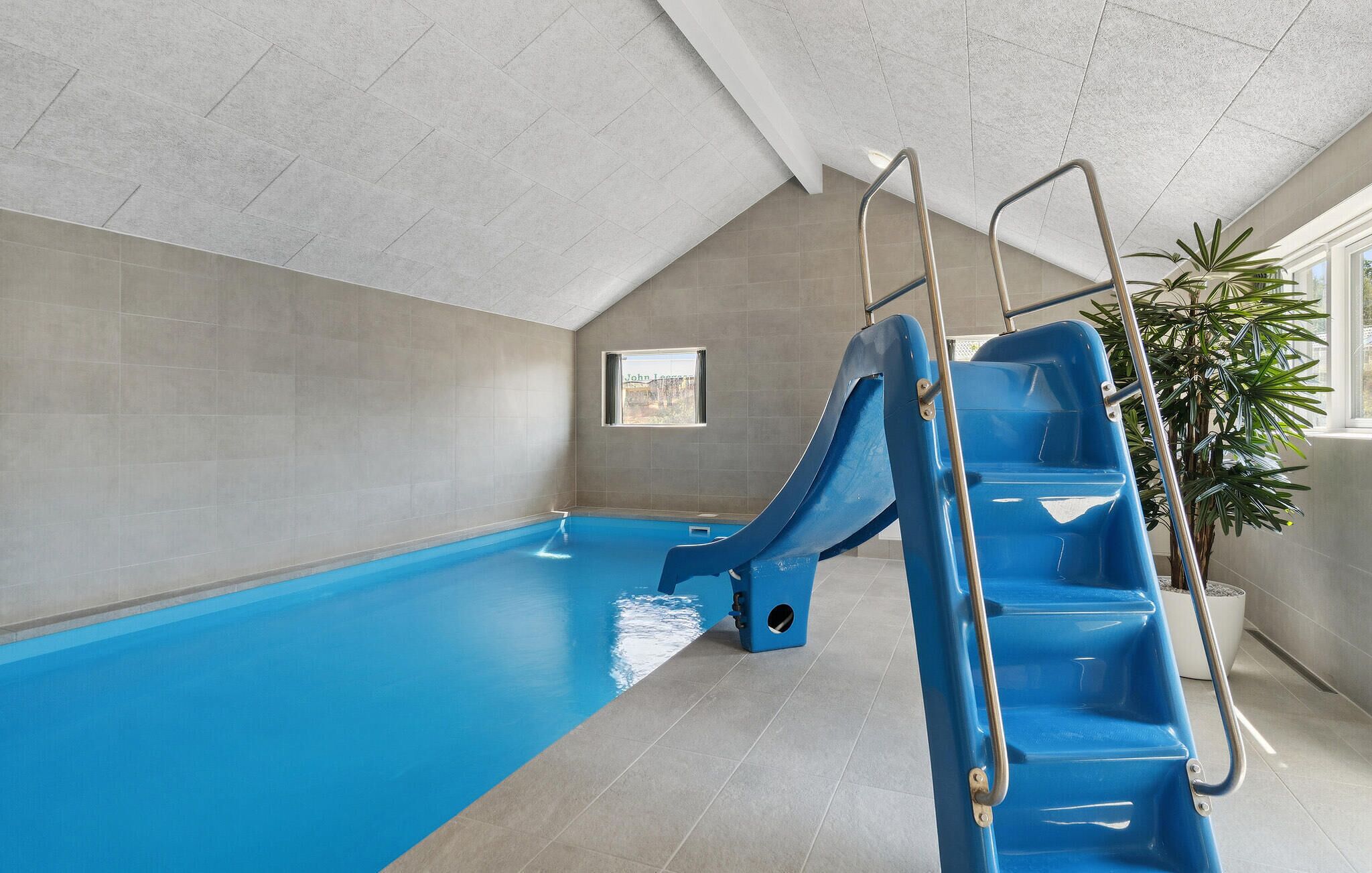 An indoor swimming pool with a blue slide, a ladder, and a potted plant.