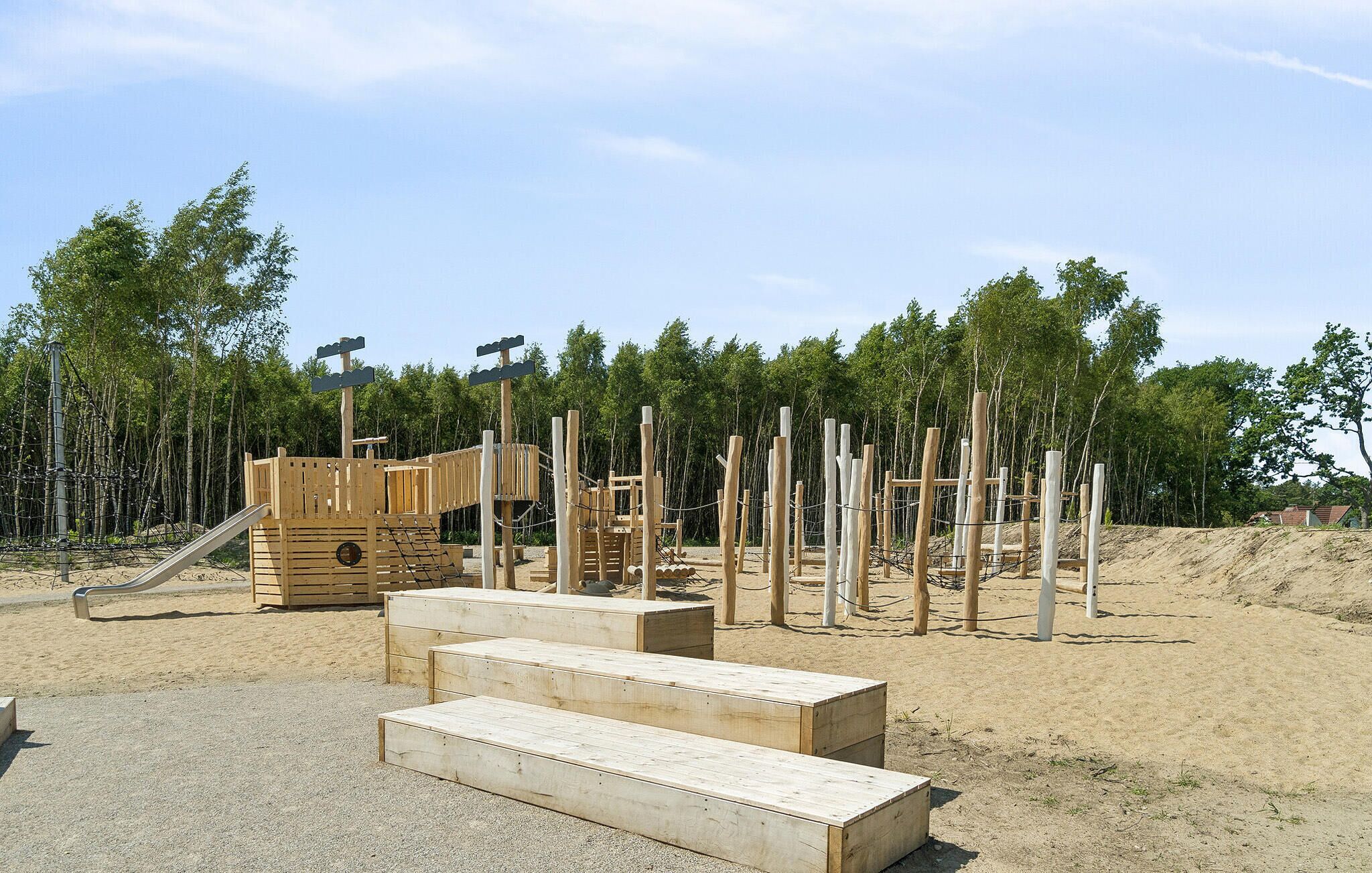 A playground with wooden structures and a slide, surrounded by trees and sandy ground.