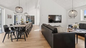 A modern living room with a large TV, black sofas, and a dining area with a long table and chairs. - Gorgeous home in Glesborg with sauna (Glesborg)