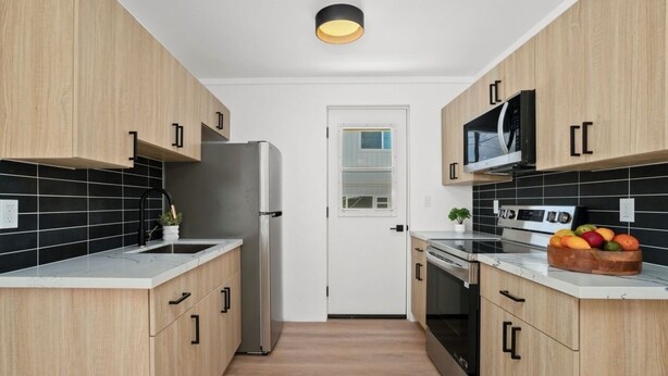 Een moderne keuken met houten kastjes, een roestvrijstalen koelkast, een magnetron en een oven. Op het aanrecht staat een fruitschaal en bij de gootsteen een kleine plant.