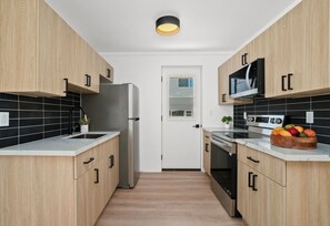 A modern kitchen with wooden cabinets, a stainless steel refrigerator, a microwave, and an oven. There is a fruit bowl on the counter and a small plant on the sink. - Charming 2-bedroom house in Waimea (Waimea)
