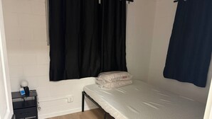 Een kleine eenpersoonskamer met een witte bakstenen muur, een zwart gordijn, een nachtkastje met een lamp en een stopcontact.