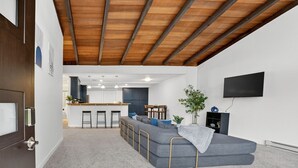 A modern living room with a grey sectional sofa, wooden ceiling beams, a kitchen in the background, and a flat-screen TV mounted on the wall. - Mid-Century Escape, Near Forest Park (Portland)